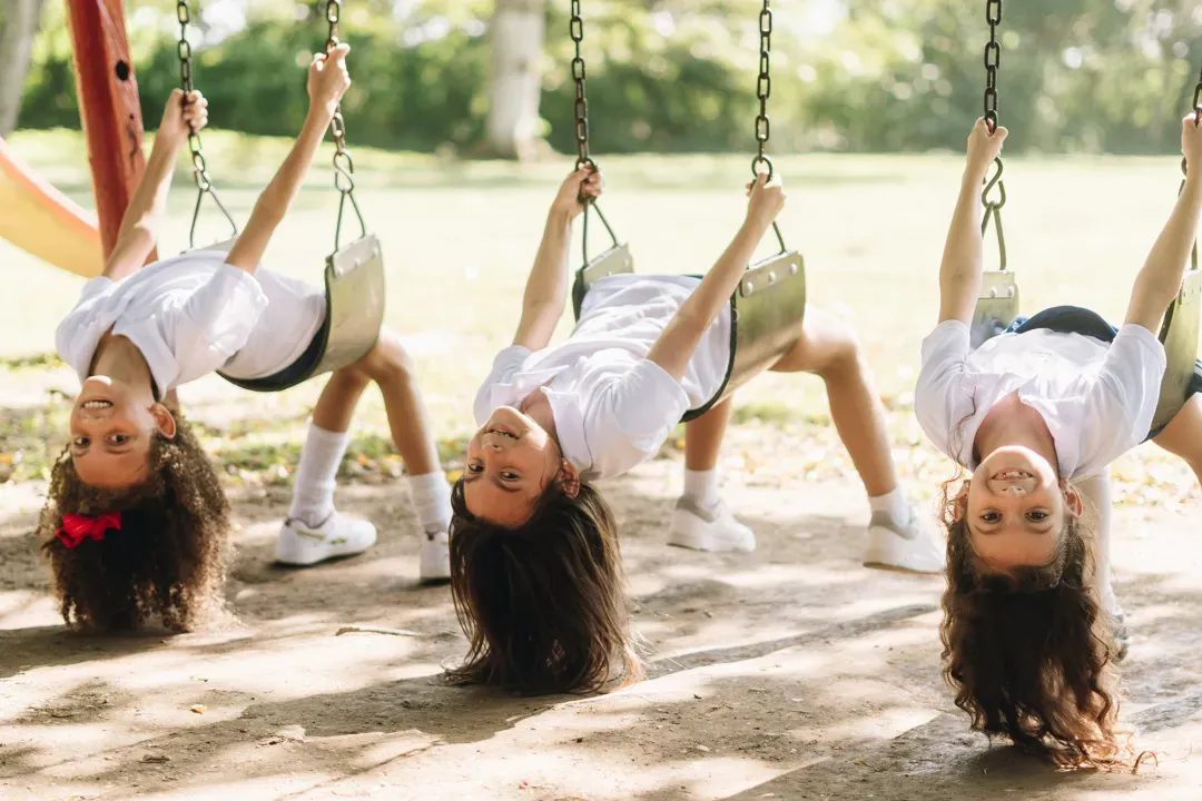 Three children hanging upside down from swings in a park, laughing